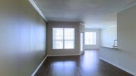 This single-floor residence offers a clean, well-appointed interior designed for comfortable living. The main living area immediately impresses with its rich, dark hardwood flooring that extends throughout, lending warmth and a polished look to the space. Crown molding lines the ceilings, adding a refined architectural touch that elevates the overall aesthetic.

Natural light is a defining feature of this home, with large windows dressed in horizontal blinds allowing ample sunlight to pour in and illuminate the open floor plan. The neutral, earthy wall tones complement the flooring and create a calm, inviting ambiance throughout.

The living area flows seamlessly toward a adjacent space — likely a dining area or kitchen — distinguished by recessed track lighting overhead and what appears to be raised countertop or bar-height surfaces, suggesting a functional and connected layout ideal for entertaining or everyday living.

The space is characterized by clean lines, understated finishes, and a cohesive design palette. White baseboards provide crisp contrast against the darker floors, while the open sightlines between rooms enhance the sense of spaciousness. Overall, the residence presents a bright, move-in-ready interior with thoughtful details throughout.