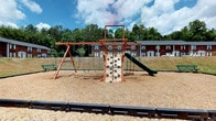 This community playground offers a welcoming outdoor recreational space designed for active play. At its center stands a well-appointed wooden play structure finished in a warm reddish-brown tone, featuring multiple activity stations including a rock climbing wall with colorful hand grips, a green slide, a swing set with two seats, and an overhead ladder traverse. The play area is surfaced with wood chips, providing a cushioned and safe ground covering, and is neatly bordered by black rubber edging. A bench is positioned nearby, offering a comfortable spot for observers. The surrounding landscape features a generous open lawn area backed by a lush treeline, creating a naturally serene environment. Brick residential buildings are visible in the background, situating the playground within an established community setting. Interior spaces associated with the property feature neutral-toned, carpeted rooms with light gray walls, white trim, and standard windows fitted with horizontal blinds that allow natural light to filter in. The rooms present a clean, move-in ready condition with a straightforward and functional layout, offering residents a comfortable and well-maintained living environment complemented by the vibrant outdoor amenity just steps away.