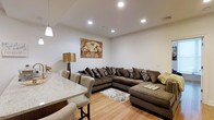 This single-floor residence features one bedroom, one bathroom, and a dedicated study, making it a versatile and functional living space. The open-concept layout connects the living and dining areas, creating a cohesive flow throughout the home. The living area is well-proportioned, offering ample room for generous seating arrangements, as evidenced by the large sectional sofa that anchors the space. Recessed lighting throughout the ceiling provides consistent, even illumination, complementing the warm tones of the light wood flooring that runs across the main living area. The dining section sits adjacent to the living room and features a marble-topped counter or bar-height surface paired with upholstered side chairs, adding a sophisticated touch. The neutral wall palette keeps the atmosphere bright and open, while the recessed lighting enhances the clean, modern aesthetic. The study offers additional flexible space suitable for a home office or guest room. Overall, the residence presents a well-designed, single-level layout that balances practicality with a polished, contemporary ambiance.