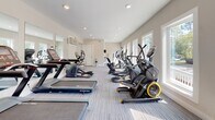 Located at 2074 Forest Hill Rd in Macon, Georgia, this fitness center offers a well-equipped, single-floor workout facility designed for comfort and functionality. The space features light-toned walls, recessed lighting, and large white-framed windows along multiple walls that flood the interior with natural light and provide pleasant views of the surrounding tree-lined grounds.

The flooring throughout consists of durable, striped carpet tiles in neutral tones, providing a practical and clean surface for exercise. The room is generously proportioned, with a long, open layout that comfortably accommodates a broad range of cardio machines, including treadmills, ellipticals, a StairMaster, and stationary bikes from brands such as Star Trac and Precor. A multi-functional cable and weight training station is also present, catering to strength training needs.

Large wall-mounted mirrors enhance the sense of space and allow users to monitor their form. A wall-mounted HVAC unit ensures climate control year-round. A seating area with chairs provides a convenient rest space. The facility includes clearly marked exit doors, offering easy access and safety compliance. Overall, the space presents a bright, airy, and fully functional fitness environment suitable for a wide range of exercise routines.