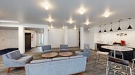 Located at 3601 Midland Ave in Memphis, Tennessee, this contemporary community living room offers a well-appointed, single-floor space designed for both relaxation and collaboration. The open-plan layout is anchored by a comfortable lounge area furnished with mid-century modern sofas and accent chairs in neutral grey tones, arranged around round wooden coffee tables atop a textured area rug — creating an inviting space for socializing or unwinding.

Adjacent to the lounge is a defined conference and dining area, featuring a long communal table with modern seating, a wall-mounted flat-screen display, and a stylish pendant light fixture overhead — ideal for meetings or group gatherings. A built-in kitchenette with dark cabinetry, a refrigerator, and pendant lighting rounds out the amenities, offering convenient access to refreshments.

The space is finished with a combination of tile and carpet flooring, and bright white walls that enhance the clean, modern aesthetic. Recessed and flush-mount ceiling lighting illuminates the interior evenly throughout. Large sliding glass doors along one wall flood the space with natural light and provide seamless access to an outdoor patio area, offering views of the surrounding property. Structural columns add architectural definition while maintaining the open, airy feel of the room.