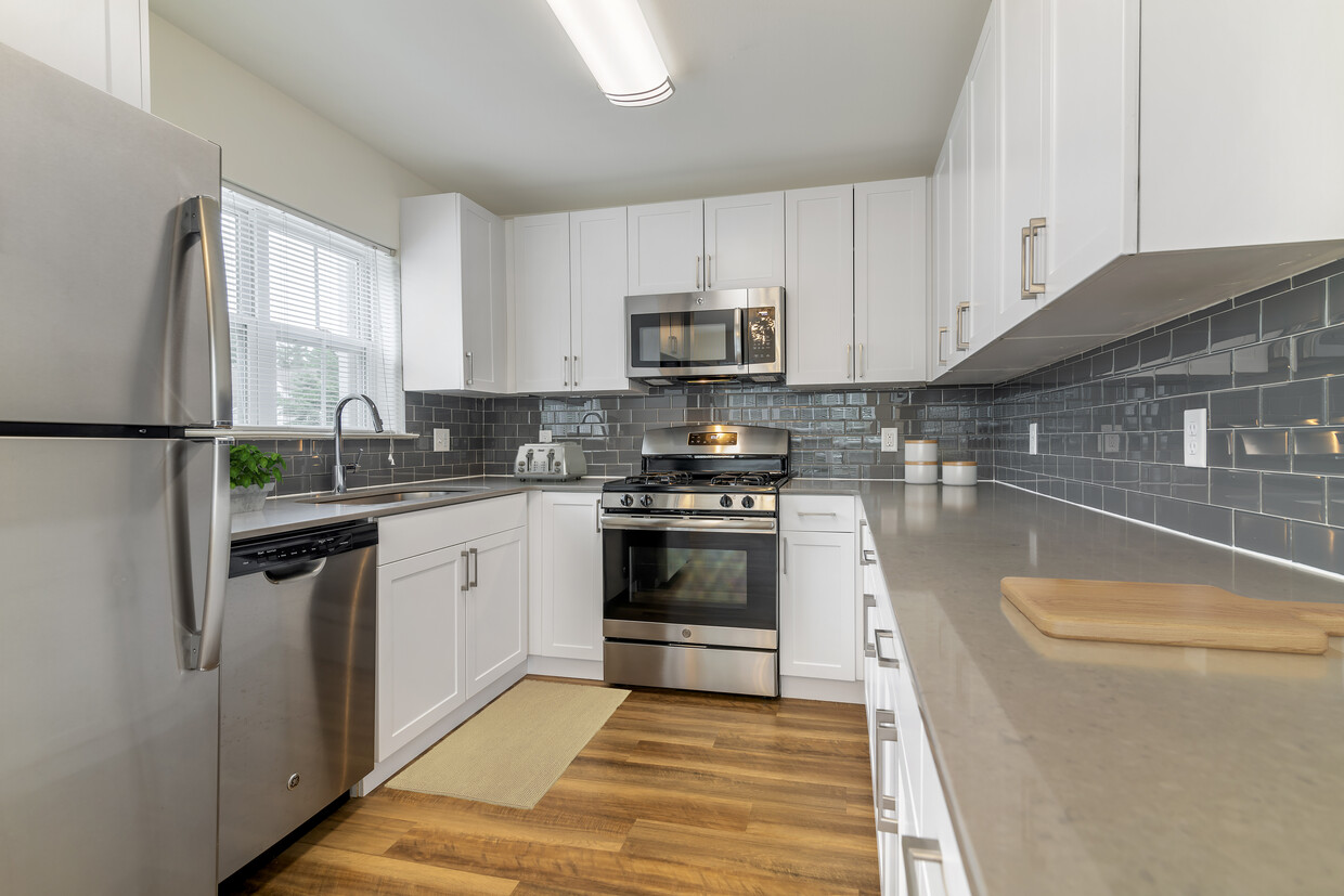 Cocina renovada del Paquete II con gabinetes blancos, encimeras de cuarzo gris, salpicadero de azulejos grises, electrodomésticos de acero inoxidable y pisos de madera de superficie dura en todas las áreas comunes. Armarios de cierre suave y microondas disponibles en determinados hogares - eaves Wilmington and Wilmington West