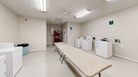 Located at 2300 Sherry Circle in Milledgeville, Georgia, this community laundry facility offers a clean, functional, and well-organized space designed to meet residents' everyday laundry needs. The single-floor layout provides a comfortable and efficient environment with a straightforward, open floor plan.

The laundry room is equipped with multiple top-loading and front-loading washing machines, paired with a row of matching top-loading dryers along one wall, providing ample capacity for simultaneous use. Two large folding tables are positioned centrally in the room, offering generous surface area for sorting and folding clothes with ease.

Bright fluorescent lighting illuminates the entire space evenly, and a ceiling fan provides additional air circulation, keeping the room comfortable during use. The neutral-toned walls and light-colored tile flooring contribute to a clean and welcoming atmosphere.

A beverage vending machine is conveniently located near the entrance, adding a practical amenity for residents. Additional utility connections are visible along the walls, indicating flexibility for future equipment as needed. A private restroom is accessible from within the facility, further enhancing convenience.

Overall, this laundry facility is a well-maintained, practical shared space that prioritizes functionality and ease of use for its community residents.