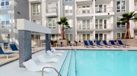 This outdoor amenity space is designed to deliver a resort-like atmosphere within a residential setting. The centerpiece is a generously sized swimming pool with clear, blue water, surrounded by a spacious pool deck furnished with a mix of white lounge chairs and navy blue seating, providing ample space for relaxation and recreation. A palm tree adds a tropical accent near the pool's edge, complementing the modern multi-story building facade featuring neutral grey siding, large windows, and private balconies that overlook the pool area.

A dedicated spa and hot tub area offers a quieter retreat, screened by neatly trimmed hedges and mature trees that create a sense of privacy. Lounge chairs are positioned poolside to take advantage of the open-air setting. The landscaping throughout is lush and well-maintained, with dense shrubs, ferns, ornamental grasses, and established trees framing the walkways and pool perimeter. Concrete pathways connect the various outdoor zones, while shaded seating areas with umbrellas provide relief from direct sunlight. The overall ambiance is one of carefully curated outdoor living, balancing modern design aesthetics with natural greenery.