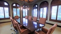 This sophisticated single-floor space is anchored by a stately boardroom that exudes a warm, Mediterranean-inspired aesthetic. The centerpiece of the room is a large, dark-stained hardwood conference table surrounded by cushioned upholstered chairs in a rich earth-tone fabric, comfortably seating a full complement of attendees. Above, a wrought-iron chandelier with ornate detailing adds a dramatic focal point and reinforces the space's classical character.

The architectural design is particularly distinguished by a series of tall, arched windows framed in dark wood, fitted with plantation shutters that allow for precise control of natural light. These arched windows lend the room a sense of grandeur and height, while offering glimpses of lush greenery outside. The walls are finished in a neutral warm tone that complements the deep wood accents throughout.

A coffered or tray ceiling detail crowns the room, further elevating its refined ambiance. The overall atmosphere balances professional functionality with architectural elegance, making the space well-suited for high-level meetings, executive gatherings, or formal presentations. The consistent use of dark wood tones, soft upholstery, and classical architectural elements creates a cohesive and impressive environment.