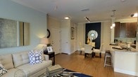 This single-floor residence offers a thoughtfully designed open-concept layout that seamlessly connects the living and dining areas, creating a cohesive and inviting atmosphere. The space is anchored by warm hardwood flooring that runs throughout, complementing the neutral, light-toned walls that lend an airy and welcoming feel.

The living area is furnished with a cream-toned sofa accented by patterned throw pillows, a sleek dark console table with a table lamp, and a decorative mirror, contributing to a balanced blend of comfort and style. Crown molding details add an elegant architectural touch along the ceiling line.

The dining area stands out with a striking navy blue accent wall that serves as a bold focal point. A circular decorative mirror with an ornate frame is mounted centrally on the accent wall, flanked by a gold decorative vase, adding a touch of glamour. The dining set features cream upholstered chairs and a dark table, creating a refined contrast.

Track lighting above the dining space and a flush-mount ceiling light near the entry ensure well-distributed illumination throughout. White interior doors and clean trim details reinforce the polished, classic aesthetic of the overall space.