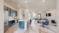 This single-floor communal space is designed with a clean, contemporary aesthetic that balances functionality and comfort. The open layout connects a well-appointed kitchen area to a spacious dining and lounge zone, creating a seamless flow throughout.

The kitchen is a standout feature, outfitted with bold teal shaker-style cabinetry, white quartz countertops, a subway tile backsplash, and stainless steel appliances including a full-size refrigerator. A central island with a built-in sink and bar seating provides an additional gathering point and casual workspace.

The dining area accommodates multiple groups simultaneously, furnished with marble-topped tables and sleek navy chairs arranged across light wood-look flooring. Recessed lighting throughout ensures consistent illumination, while large windows along one wall flood the space with natural light and frame views of an outdoor courtyard. Tall potted palms positioned near the windows add a lively, organic touch to the otherwise neutral palette.

A dedicated media zone features wall-mounted flat-screen televisions, making the space well-suited for presentations or group viewing. Direct exterior access through glazed doors connects the interior to the surrounding outdoor environment, enhancing the bright, airy ambiance of the overall space.