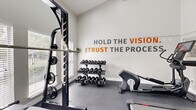 Located at 400 Putnam Dr in Tallahassee, FL, this dedicated fitness room delivers a comprehensive workout environment within a compact, efficiently designed single-level space. The room features soaring ceilings that lend a sense of openness, complemented by large windows fitted with white blinds that invite natural light and offer views of an outdoor pool area and surrounding greenery.

The flooring consists of durable dark rubber gym tiles, providing both safety and functionality throughout. A full wall of floor-to-ceiling mirrors visually expands the space while allowing users to monitor their form during exercise. An eye-catching motivational wall graphic reading "HOLD THE VISION. TRUST THE PROCESS." in bold gray and orange lettering sets an energizing tone.

Strength training is well supported with a heavy-duty power rack, an Olympic barbell with weight plates, a weight bench, and a multi-tiered dumbbell rack stocked with a comprehensive range of free weights. Cardio equipment includes a treadmill, an elliptical trainer, and a stationary bike, each outfitted with interactive touchscreen displays. The room also connects to adjacent living areas through glass panel openings, maintaining a sense of integration with the broader property while keeping the fitness area clearly defined.