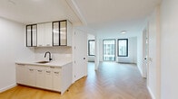 Located at 163 Willoughby Street in the heart of Downtown Brooklyn, this thoughtfully designed two-bedroom, two-bathroom residence offers a clean, modern aesthetic across a single-level floor plan. Warm herringbone-patterned hardwood floors flow throughout the living areas and bedrooms, lending a refined elegance to the bright, white-walled interiors. Both bedrooms feature large, black-framed floor-to-ceiling windows that fill the rooms with natural light and offer views of the surrounding Brooklyn skyline. The primary bedroom includes a double-door closet with matte black hardware, providing generous storage. Each bedroom is equipped with individual climate control units for personalized comfort. The two bathrooms are spa-inspired and distinctly designed. One features soft beige mosaic tile walls surrounding a soaking tub with a glass panel enclosure, a wood-paneled floating vanity with open shelving, and built-in wall niches. The second bathroom showcases gray stone-look wall tile, a terrazzo-style floor, a wood floating vanity with black metal legs, and an illuminated mirror framed by warm pendant lighting. A connecting hallway ties the residence together, offering glimpses into the kitchen and living areas beyond, completing this polished urban home.