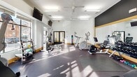 This fitness center offers a thoughtfully designed, single-floor layout that provides a comfortable and functional environment for a full range of workouts. The space is organized to maximize usability, with distinct zones for cardio, strength training, and free-weight exercises.

The main workout area features high ceilings that enhance the open, airy feel of the room. Overhead ceiling fans and flush-mounted lighting ensure consistent airflow and visibility throughout. Durable rubber flooring covers the workout zones, providing both safety and comfort underfoot.

Large windows along one wall flood the space with natural light and offer views of the surrounding neighborhood, creating an inviting atmosphere. Wall-mounted flat-screen televisions are positioned throughout for entertainment during workouts.

The equipment selection is comprehensive, including elliptical trainers, a stationary bike, a rowing machine, and a cable crossover strength machine. A rack of colorful medicine balls is conveniently located near the entrance for functional training exercises. A full-length mirror along one wall supports form monitoring and visually expands the space.

Access is provided through a set of glass double doors, contributing to the bright, welcoming character of the facility. The clean white walls with warm wood-toned baseboards give the space a crisp, professional aesthetic suited to an active lifestyle.
