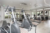 Located at 3385 Atlanta Rd SE in Smyrna, Georgia, this well-appointed fitness center is designed to support a comprehensive workout experience across two distinct zones. The space is defined by bold orange accent walls, a striking curved ceiling detail with integrated LED strip lighting, and a clean white-and-gray color palette that creates an energetic yet focused atmosphere throughout.

The strength and functional training area features a full rack of dumbbells, medicine ball storage, kettlebells, a workout mat, stability balls, and multiple Cybex cable and weight machines. Floor-to-ceiling mirrors line one wall, enhancing the sense of spaciousness. Recessed lighting and a ceiling fan ensure comfort during intense training sessions.

The dedicated cardio zone is bathed in natural light, courtesy of expansive floor-to-ceiling glass doors and large transom windows that offer views of lush outdoor greenery. This section houses a row of treadmills, elliptical trainers, and stationary bikes, all arranged on a warm wood-toned flooring surface that contrasts with the rubber sport flooring in the strength area. A ceiling fan and mounted televisions add to the comfort and entertainment options.

Together, the two zones provide a fully equipped, single-floor fitness facility suited for a wide range of training needs.