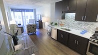 This single-floor unit presents an open-concept layout that seamlessly integrates the kitchen and living area into one cohesive space. The kitchen is appointed with dark espresso cabinetry, both upper and lower, providing ample storage. A stainless steel sink and dishwasher lend a contemporary edge, while the mosaic tile backsplash in soft greens and grays adds a decorative focal point. Light-toned countertops contrast neatly against the dark cabinetry.

Warm, wide-plank hardwood flooring extends throughout the entire space, creating visual continuity and a sense of depth. The living area is thoughtfully arranged to maximize the compact footprint, with seating positioned to take advantage of the large floor-to-ceiling windows at the far end. These windows allow generous natural light to flood the room, enhancing the open and airy ambiance.

Exposed concrete ceiling elements add an urban, industrial character to the space, balancing the warmth of the wood floors. Overall, the unit offers a modern, functional aesthetic suited to efficient, stylish urban living.
