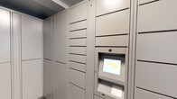 The Parcel Room is a multifunctional space that combines practical package management with an inviting communal atmosphere. At its core, the parcel area features a bank of modern, floor-to-ceiling smart lockers finished in a neutral gray. These secure units are equipped with a digital touchscreen terminal and keypad, allowing residents to retrieve deliveries conveniently at any time.

Adjacent to the parcel area, a sophisticated lounge space extends the room's functionality. The lounge is decorated with an elegant floral-patterned area rug in warm earth tones, anchoring multiple seating arrangements with white upholstered dining chairs surrounding dark wood round tables. A striking stone fireplace serves as a focal point against a teal accent wall, adding warmth and visual interest.

Large windows line the perimeter, flooding the space with natural light and offering views of the surrounding outdoor greenery. Decorative touches include wall-mounted white ceramic planters with cascading succulents and trailing greenery, as well as potted palms placed throughout. Recessed lighting and a statement chandelier complete the refined, welcoming ambiance of this well-designed communal space.