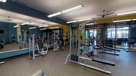 This fitness center offers a comprehensive workout environment designed to support a wide range of exercise routines. The space is outfitted with multiple Life Fitness strength and resistance training machines, including a cable pulley system, leg press, and various selectorized weight stations, providing users with ample options for full-body training. The flooring is finished with durable rubber gym matting, which offers both safety and comfort during workouts. Large floor-to-ceiling mirrors line one wall, enhancing the sense of space and allowing users to monitor their form during exercise. The interior color scheme features bold teal and yellow accent walls, giving the space a modern and energetic ambiance. Overhead, industrial-style fluorescent strip lighting ensures the area is well-lit throughout, while ceiling fans contribute to air circulation and comfort. A glass partition or window wall on one side of the room allows natural light to filter in and provides a visual connection to an adjacent area. The overall layout is open and organized, allowing for easy navigation between equipment. This fitness center is thoughtfully designed to meet the needs of a diverse range of users seeking a functional and motivating exercise environment.