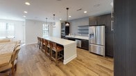 Located in Cedarburg, Wisconsin, this thoughtfully designed single-floor space offers an open-concept layout that seamlessly connects its key living areas. The interior is finished with warm wood-look flooring throughout, neutral-toned walls, and recessed lighting that creates a bright, welcoming ambiance.

The living area is generously proportioned, featuring large windows dressed with horizontal blinds that allow abundant natural light. A statement ring chandelier with warm brass accents serves as a focal point, complemented by a mix of rich brown leather sofas and neutral upholstered seating arranged around a marble-top coffee table. A wall-mounted flat-screen TV adds a modern touch.

The kitchen is a standout feature, boasting dark cabinetry with sleek hardware, crisp white quartz countertops, and a full suite of stainless steel appliances. Open shelving with a tile backsplash adds visual interest, while a large island with bar seating and pendant lighting makes it ideal for both cooking and casual gathering.

The dining area accommodates a large natural wood farmhouse table with ample seating, flanked by an elegant wall with white decorative panel molding — adding a classic architectural detail to the space. Views to the surrounding streetscape are available through the multiple windows, connecting the interior to the charming Cedarburg neighborhood outside.