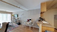 This single-floor space is defined by its open-concept layout, seamlessly connecting the kitchen, dining, and living areas into one cohesive environment. The vaulted ceilings are a standout architectural feature, adding a sense of height and airiness that enhances the overall spaciousness of the interior. A track lighting system runs along the ceiling, providing adjustable illumination throughout the main living area.

The kitchen is positioned along one side, featuring warm honey-toned wood cabinetry, granite-style countertops, and a peninsula that serves as a natural divider between the kitchen and dining space. This peninsula also functions as a casual bar or prep area.

The dining area sits centrally within the open plan, accommodating a modest white table and chairs. The living area extends toward the far end of the space, anchored by hardwood-style light oak flooring. A low-profile shelving unit lines one wall, offering storage and display space alongside a wall-mounted television. Neutral gray walls throughout provide a clean, understated backdrop that complements the warm tones of the cabinetry and flooring.