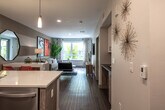 This contemporary single-floor residence showcases a thoughtfully designed open-concept layout that seamlessly connects the kitchen, dining, and living areas. Rich wood-look flooring flows throughout, complementing the clean, modern aesthetic.

The kitchen is a standout feature, equipped with stainless steel appliances including a refrigerator, range, and microwave. White gloss upper cabinetry is paired with warm wood-toned lower cabinets, while a mosaic tile backsplash adds texture and visual interest. A peninsula with a white quartz countertop and bar seating provides both additional workspace and a casual dining option. Pendant lighting above the peninsula enhances the kitchen's contemporary feel.

The open living area offers ample room for relaxation and entertaining, with natural light streaming in through large windows. The space features neutral gray walls accented with bold artwork, adding a modern, curated character to the room.

A louvered-door closet and additional storage solutions contribute to the unit's functional design. The bathroom features a bold red accent wall, a wood-toned vanity with an undermount sink, a large mirror, and a tub-shower combination, blending practicality with a striking visual style.

Overall, this residence delivers a cohesive modern design with quality finishes and a versatile, open floor plan.