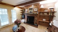 Located at 9696 Hayne Blvd in the Pontchartrain Oaks neighborhood of New Orleans, this single-story home exudes a warm and inviting character defined by its rich wood-paneled walls and exposed wooden ceiling beams. The living area serves as the heart of the home, centered around a striking wood-surround fireplace with a dark hearth and decorative mantel, flanked by built-in shelving units that provide both display and storage space. The diagonal wood paneling surrounding the fireplace adds a distinctive visual texture, giving the room a cozy, lodge-inspired aesthetic. Natural light filters in through windows fitted with blinds, complementing the warm tones of the interior woodwork. Open shelving along one wall offers additional functional display space, while carpeted flooring adds to the comfortable, lived-in feel of the room. The overall ambiance of the home is one of classic, rustic charm with a welcoming atmosphere, reflecting the character and tradition often found in established New Orleans neighborhoods. The single-floor layout ensures easy accessibility and a cohesive flow throughout the property.