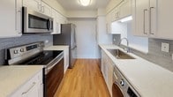 This single-floor unit offers a thoughtfully organized layout with consistent warm-toned hardwood flooring throughout, complemented by light grey walls and white trim that create a clean, neutral aesthetic. The open living and dining area is generously proportioned, featuring wall sconces, a decorative chandelier, and a textured ceiling. Large windows allow natural light to filter in, framing views of the surrounding urban landscape and greenery.

The galley-style kitchen is well-equipped with white shaker cabinetry, light quartz countertops, a grey tile backsplash, and a full suite of stainless steel appliances including a range, microwave, and refrigerator. An in-unit washer is conveniently integrated beneath the counter near the sink, adding practical value.

The unit includes two bedrooms, each finished with the same hardwood flooring and neutral palette. One bedroom features a spacious walk-in closet outfitted with wire shelving, while the other enjoys direct access to natural light through a large window with city views. Both rooms include wall sconce lighting.

The full bathroom is finished with classic white square tile on the floors and tub surround, complemented by a warm wood-toned vanity and chrome fixtures, providing a clean and functional design.