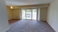 This single-floor residence offers a welcoming and functional layout designed for comfortable everyday living. The main living area is a standout feature, boasting wall-to-wall carpeting that adds warmth underfoot and a sense of coziness throughout the space. Large sliding glass doors span nearly the full width of the room, flooding the interior with abundant natural light and providing direct access to a private balcony. The balcony offers pleasant views of mature surrounding trees, creating a serene, nature-connected ambiance. Sheer white vertical blinds frame the glass doors elegantly, allowing for flexible light control and privacy. A ceiling fan is positioned overhead, contributing to year-round comfort and air circulation. The neutral, light-toned walls serve as a versatile backdrop suitable for a variety of interior styles. A floor-level vent indicates a central HVAC system for climate control throughout the unit. The overall atmosphere of the space is bright, open, and tranquil, with the natural wooded backdrop visible through the expansive glazing serving as a defining characteristic of the living environment.