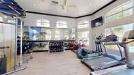 Located in Phoenix, AZ, this well-equipped fitness center offers a comprehensive workout environment within a bright, airy single-room layout. The space features soaring ceilings with decorative crown molding, large arched windows along multiple walls, and ceiling fans that promote air circulation and comfort during exercise. Natural light floods the interior, with views of an outdoor pool area visible through the expansive windows, creating an inviting and motivating atmosphere.

The exercise room is outfitted with a full lineup of cardio equipment, including multiple treadmills, elliptical trainers, a stair climber, and a row of red spin bikes. A dedicated free weights section lines one wall, complete with a tiered dumbbell rack and an adjustable weight bench, all backed by full-length mirrors that enhance the sense of space. A cable resistance machine provides additional strength training options. Wall-mounted flat-screen TVs are positioned throughout the room for entertainment during workouts.

The light wood-toned flooring contributes to a clean, modern aesthetic, while a bold blue graphic accent wall adds a dynamic visual element. A glass door provides direct access to the outdoor area. The overall design balances functionality and style, making this fitness center a practical and well-appointed space for a full range of exercise routines.
