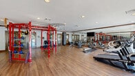 The Lofts Hillside Fitness Center at 1315 Creekshire Way in Winston-Salem, NC, is a thoughtfully designed exercise facility occupying a single floor. The space features warm hardwood-style flooring throughout, recessed lighting, and crown molding details that lend a polished, upscale feel uncommon in typical fitness facilities.

The exercise room is generously proportioned, offering ample open floor space to accommodate a wide variety of workout styles. A bold red multi-functional training rig serves as a centerpiece, equipped with TRX suspension straps, battle ropes, and a heavy punching bag, supporting functional and high-intensity training. Overhead, a curved cable pulley system with gymnastic rings adds further versatility.

Along one wall, floor-to-ceiling mirrors visually expand the space and are accompanied by a wall-mounted flat-screen television. Large windows along the perimeter flood the room with natural light and provide views of an outdoor area featuring a covered gazebo. The cardio zone is well-stocked with treadmills, rowing machines, and stationary bikes, while a dedicated strength area houses a Smith machine, weight bench, and additional free weights.

Wood-framed glass doors provide a refined entry to the facility. Fitness center rules are clearly posted, and amenities such as hand sanitizer dispensers and waste bins are conveniently placed. The overall ambiance is energetic yet refined, making it a fully functional and inviting space for residents.