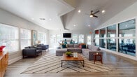 This single-floor community clubhouse is designed with a clean, contemporary aesthetic and a welcoming mid-century modern flair. The expansive main lounge serves as the centerpiece of the space, featuring soaring vaulted ceilings with a geometric coffered detail, a ceiling fan, and abundant recessed lighting. Large windows on multiple walls flood the room with natural light and offer views of the surrounding community. The lounge is anchored by a generously sized sectional sofa atop a chevron-patterned area rug, complemented by a walnut coffee table and coordinating accent chairs — all set over light wood-tone flooring that runs throughout the space.

A dedicated seating nook near the entry provides a quieter retreat, furnished with a pair of upholstered arm chairs, vibrant abstract wall art, and a wall-mounted display screen. Adjacent to the lounge, a high-top dining table with teal accent chairs offers a casual gathering spot beneath a wall-mounted TV, surrounded by windows that allow natural light to pour in.

The kitchen area features gray shaker-style cabinetry, white countertops, stainless steel appliances, and a breakfast bar with leather counter stools — ideal for casual dining or entertaining. A fitness center with cardio equipment and exercise accessories is conveniently visible through interior windows, adding a functional amenity to this well-appointed community hub.