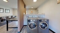 Located at 199 Aegean Way in Vacaville, California, this single-floor community laundry facility is designed with both functionality and comfort in mind. The space is outfitted with a row of commercial-grade front-loading washers and dryers finished in sleek stainless steel, providing ample capacity for residents. The machines are positioned along the walls with generous counter space above, ideal for sorting and staging laundry. The flooring throughout features a wood-look vinyl plank in a light, striped tone that adds a contemporary feel to the utilitarian space. A dedicated folding area is furnished with a large dark-surfaced table and a wooden bench, offering convenient workspace for residents. An additional counter-height granite-topped bar runs along a large window, accompanied by industrial-style stools — a thoughtful touch that allows residents to wait comfortably while enjoying views of the surrounding landscaped grounds and mature trees. The walls are painted in neutral tones accented by a warm taupe feature wall, and framed artwork is displayed throughout, elevating the ambiance beyond a typical laundry room. Natural light filters in through multiple windows, creating a bright and welcoming atmosphere. The space is clean, well-maintained, and thoughtfully appointed for everyday community use.