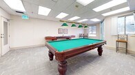 This well-appointed game room offers a comfortable and engaging recreational space designed for leisure and social activity. The centerpiece of the room is a full-size pool table with a rich mahogany finish and classic green felt surface, illuminated overhead by a brass-accented pendant light fixture fitted with green shades that casts focused light across the playing surface. A dedicated cue rack mounted on the wall keeps equipment neatly organized and accessible.

Complementing the pool table is a full-length shuffleboard table positioned along one wall, providing an additional layer of entertainment options. A set of tall bistro-style tables and chairs occupy a corner of the room, creating a casual seating area ideal for spectators or social gatherings. Framed artwork and informational displays decorate the neutral-toned walls, adding a polished, welcoming feel to the space.

The room is finished with patterned low-pile carpeting throughout, contributing to both comfort underfoot and sound absorption. A drop-tile ceiling with recessed panel lighting ensures bright, even illumination across the entire floor area. Windows along one wall allow natural light to filter into the space, enhancing the open and airy atmosphere. A secondary windowed alcove and an exterior-facing door further connect the interior to its surroundings, rounding out this versatile and inviting recreational amenity.