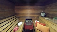 This sauna is a thoughtfully appointed wellness space encased entirely in rich, dark-toned horizontal wood planking that lines the walls, ceiling, and floor, creating a warm and enveloping atmosphere. At the heart of the room stands a central electric sauna heater topped with heat-retaining stones, serving as the functional and visual focal point of the space. Tiered wooden benches line both sides of the room, offering multiple seating and reclining positions to accommodate varying heat preferences, as temperatures naturally rise toward the upper levels. The slatted bench construction allows for airflow and comfort during extended sessions. Thoughtful touches elevate the experience, including decorative cushions in warm coral and peach tones, folded white towels, a wire basket stocked with sauna amenities, a loofah, and a small potted succulent that adds a natural accent. A doorway connects the sauna to an adjoining anteroom finished with light tile flooring, providing a cool-down area and transition space. The overall ambiance is intimate, grounded, and restorative, with the natural wood tones contributing to a traditional sauna aesthetic.