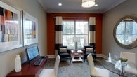 This thoughtfully designed single-floor space makes a strong visual impression through its careful use of color, texture, and layout. The lounge area serves as a central focal point, anchored by a rich burnt-orange accent wall that contrasts elegantly against the warm neutral tones of the surrounding walls and crown molding. Bold black-and-white striped curtains frame a large window that invites generous natural light and offers views of the surrounding outdoor area, enhancing the room's open and airy feel. Two sleek black armchairs with decorative accent pillows flank a glass-top coffee table, creating an intimate seating arrangement well-suited for conversation or relaxation. A drum pendant light fixture overhead adds a modern, polished touch to the space. Near the entrance, a rich wood media console supports a monitor and keyboard, suggesting a functional workspace component integrated seamlessly into the room. White contemporary chairs add further seating options, contributing to the space's versatile, multi-use character. A framed piece of colorful wall art provides an additional layer of visual interest. The combination of patterned carpeting, refined trim work, and purposeful furnishings gives the overall space a professional yet welcoming ambiance.