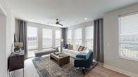This thoughtfully designed single-floor residence offers a cohesive blend of modern aesthetics and functional living. The open-concept living area is bathed in natural light, courtesy of a generous array of large windows dressed with horizontal blinds and elegant grommet-top curtains. A ceiling fan anchors the space, which features light hardwood-style flooring and a neutral color palette that lends a clean, contemporary feel.

The kitchen is a standout feature, outfitted with white shaker-style cabinetry, light quartz countertops, a subtle tile backsplash, and a full suite of stainless steel appliances. A central island with bar seating and pendant lighting above makes it ideal for both cooking and casual dining. Adjacent to the kitchen, the dining area provides a dedicated space for seated meals, with a large window offering natural light.

The bedrooms are carpeted for comfort, each equipped with a ceiling fan, ample natural light from well-placed windows, and a calm, neutral tone. The primary bath features a wide vanity with a large mirror, while a secondary full bath includes a tub-shower combination with tile detailing. An additional half bath offers a single vanity and a frosted privacy window. Light wood-style flooring runs consistently throughout the non-carpeted areas, tying the space together seamlessly.