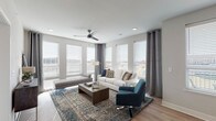 This thoughtfully designed single-floor residence offers a cohesive blend of modern aesthetics and functional living. The open-concept living area is bathed in natural light, courtesy of a generous array of large windows dressed with horizontal blinds and elegant grommet-top curtains. A ceiling fan anchors the space, which features light hardwood-style flooring and a neutral color palette that lends a clean, contemporary feel.

The kitchen is a standout feature, outfitted with white shaker-style cabinetry, light quartz countertops, a subtle tile backsplash, and a full suite of stainless steel appliances. A central island with bar seating and pendant lighting above makes it ideal for both cooking and casual dining. Adjacent to the kitchen, the dining area provides a dedicated space for seated meals, with a large window offering natural light.

The bedrooms are carpeted for comfort, each equipped with a ceiling fan, ample natural light from well-placed windows, and a calm, neutral tone. The primary bath features a wide vanity with a large mirror, while a secondary full bath includes a tub-shower combination with tile detailing. An additional half bath offers a single vanity and a frosted privacy window. Light wood-style flooring runs consistently throughout the non-carpeted areas, tying the space together seamlessly.