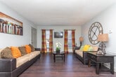 This single-floor residence offers a thoughtfully designed layout that balances comfort and functionality across its key living spaces. The kitchen is a standout feature, equipped with dark espresso cabinetry, a tile backsplash, stainless steel appliances including a range and built-in microwave, and a light-toned peninsula countertop that provides additional prep space. Rich dark hardwood-style flooring runs through the kitchen and connects seamlessly to the adjacent dining area, where a pendant light fixture illuminates a round dining table set beneath a decorative wall accent. The living room is bright and welcoming, featuring multiple windows dressed with bold plaid curtains, dark flooring, and a wall-mounted television. A large decorative clock serves as a focal point on one wall, adding character to the space. Both bedrooms are carpeted and well-lit by natural light from multiple windows, each outfitted with ceiling fans for year-round comfort. The bathroom features tile wainscoting, dark cabinetry with a light countertop, a wide vanity mirror with overhead lighting, and a tub-shower combination. Throughout the residence, textured ceilings and neutral wall tones create a cohesive, understated aesthetic that provides a versatile backdrop for a variety of interior styles.