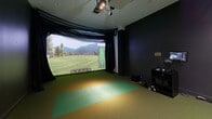 Located at 15 Farm Springs Road in Farmington, Connecticut, this dedicated indoor golf simulator room offers a fully immersive golfing experience in a compact, purpose-built space. The room is finished with dark, matte walls that minimize glare and create a focused environment, complemented by recessed ceiling lighting and professional-grade overhead spotlights that illuminate the hitting area. The centerpiece of the space is a large curved projection screen at the front of the room, displaying realistic, high-definition golf course simulations complete with shot-tracking data such as carry distance, distance to pin, and course maps. Overhead, a mounted launch monitor and projector system captures and analyzes each shot with precision. The flooring consists of turf-style carpeting, with a distinct green hitting mat positioned in front of the screen to replicate the feel of a real fairway or tee box. Black curtain panels flank the projection screen, contributing to the room's immersive atmosphere. Along the rear wall, two cushioned leather lounge chairs provide comfortable seating for players awaiting their turn or spectators. A door with an illuminated exit sign and a golf bag rack complete the functional layout, making this a well-equipped and thoughtfully designed simulator suite.