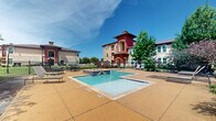 Located at 2900 Wildflower Dr in Bryan, Texas, this outdoor amenity area serves as a central gathering space within a Mediterranean-inspired residential community. The space features two distinct water elements: a generously sized rectangular swimming pool with clearly marked depth indicators and a separate spa/hot tub finished with decorative blue tile accents. Both water features are set within an expansive stamped concrete deck finished in warm earth tones, providing ample room for lounging and entertaining. Sun loungers and outdoor seating with tables are positioned around the deck, offering comfortable spots to relax in the Texas sunshine. The pool area is enclosed by a wrought iron fence with gated entry points, ensuring privacy and security. Mature trees and lush landscaping, including flowering shrubs, add natural shade and color throughout the space. The surrounding multi-story buildings showcase a distinctive architectural style with bold red and tan stucco exteriors, arched windows, and tower accents, creating an attractive backdrop for the amenity area. A low-profile clubhouse structure with dormer windows and a pitched roof is also visible adjacent to the pool, complementing the overall community aesthetic. The entire setting benefits from abundant natural light and open sky views.