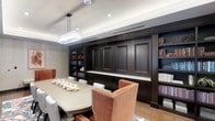 This sophisticated single-floor space centers around a strikingly designed conference or formal dining room that blends classic craftsmanship with refined contemporary sensibilities. The room is anchored by a substantial rectangular table surrounded by plush, terracotta-hued upholstered chairs with nailhead trim detailing, offering both comfort and elegance. A crystal rectangular pendant chandelier hangs overhead, casting warm light across the space and complementing the recessed lighting embedded within the tray ceiling above.

The most commanding feature of the room is the floor-to-ceiling dark espresso built-in cabinetry that spans the entire back wall. The millwork includes raised-panel cabinet doors, open display shelving stocked with books and decorative objects, and a continuous light-toned countertop surface that ties the composition together. The deep, rich tones of the cabinetry create a dramatic contrast against the lighter walls and ceiling, lending the space a sense of gravitas and formality.

Underfoot, a patterned area rug adds a touch of color and visual interest, while the overall design palette of warm neutrals, dark wood, and copper tones creates a cohesive, inviting ambiance. This space is well-suited for professional meetings, formal gatherings, or intimate dinners in a setting of distinguished style.