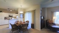 This single-floor residence showcases a thoughtfully arranged open-concept layout that connects the dining area and kitchen in a cohesive, functional design. The dining space is anchored by a dark wood table surrounded by wicker chairs, adding a warm, natural texture to the room. Above the table, a pendant light fixture with a classic silhouette provides focused, ambient illumination that complements the soft, neutral tones of the walls and carpet.

A large sliding glass door dressed with two-tone curtains in cream and brown draws the eye and allows an generous amount of natural light to filter into the space, enhancing the overall warmth and openness of the room. The carpeted flooring throughout contributes to a comfortable, quiet atmosphere.

Adjacent to the dining area, a dark wood credenza provides additional storage and display space, with a framed artwork piece mounted on the wall above it. The kitchen, visible in the background, features white cabinetry with dark accents and an overhead cabinet that extends toward the ceiling for additional storage. Track lighting along the ceiling further supports the overall illumination of the connected spaces.