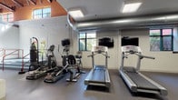 This fitness center offers a comprehensive workout environment designed to accommodate a wide range of exercise routines. The space features an industrial aesthetic, highlighted by soaring ceilings with exposed metal ductwork, structural steel columns, and a combination of white-painted brick and boldly colored accent walls in orange and gray. Natural light filters in through large windows, creating an open and energizing atmosphere.

The cardio zone is well-appointed with multiple treadmills, elliptical trainers, and a stationary bike, each equipped with individual flat-screen monitors for entertainment during workouts. A dedicated strength training area includes a cable machine with multiple attachment options, a weight rack, and a variety of free weights. Exercise balls in multiple sizes are neatly stored on a rack, supporting flexibility and core training. A standing heavy punching bag adds a boxing and functional training element to the facility.

The flooring throughout is durable rubber gym flooring, providing traction and impact absorption across all workout zones. Overhead flush-panel lighting ensures consistent illumination throughout the space. A staircase with red railings is visible, indicating access to an adjacent area. The overall layout is spacious and thoughtfully organized, allowing users to move freely between equipment zones.