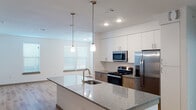 This well-appointed single-story, three-bedroom, two-bathroom residence in Leander, Texas offers a thoughtfully designed layout with modern finishes throughout. The kitchen serves as the heart of the home, showcasing a large granite-topped island with an undermount sink and pendant lighting overhead. White upper cabinetry is complemented by warm wood-toned lower cabinets, a classic subway tile backsplash, and a full suite of stainless steel appliances including a range, microwave, and french door refrigerator. Light wood-plank flooring flows seamlessly through the main living areas, enhancing the bright, open feel of the space. The primary bedroom is generously sized with direct access to an en-suite bathroom, while the additional bedrooms feature plush carpeting and ceiling fans, with natural light provided by blinded windows. The bathrooms are finished with neutral stone-tiled tub and shower surrounds and brushed nickel fixtures. A dedicated in-unit laundry room adds everyday convenience. Recessed lighting throughout keeps the interiors well-lit and clean in appearance, contributing to the home's contemporary, move-in-ready ambiance.