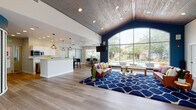 This single-story clubhouse is defined by its striking architectural design, centered around a dramatic arched window wall that floods the main lounge with natural light and frames direct views of an outdoor pool area. The vaulted ceiling in the lounge is clad in wood planking, adding warmth and texture that contrasts beautifully with the deep navy blue accent walls. Light wood-look flooring runs throughout, unifying the open-plan layout.

The lounge area is generously proportioned, accommodating a seating arrangement anchored by a bold blue patterned area rug and leather furniture. A wall-mounted television is positioned for easy viewing from the seating area.

Adjacent to the lounge, a full kitchen features white cabinetry, a large white-topped island with bar seating, stainless steel appliances, and geometric industrial-style pendant lighting overhead. Frosted glass partition walls border a separate dining area nearby, offering a degree of privacy while maintaining visual openness.

A dedicated dining nook sits beside wide windows fitted with white horizontal blinds, providing poolside views and ample natural light. The overall ambiance is modern and welcoming, balancing contemporary design elements with comfortable, community-focused furnishings.