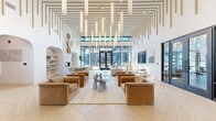 Located at 1301 Lebanon Pike in Nashville, TN, this single-floor community clubhouse delivers a refined and welcoming atmosphere through thoughtful architectural design and high-end finishes. The space is anchored by a dramatic great room with soaring double-height ceilings adorned with a striking installation of cascading cylindrical pendant lights and linear wood slat detailing — a defining design feature visible throughout. Light oak herringbone flooring flows seamlessly across the entire space, reinforcing the warm, modern aesthetic.

The main lounge area features generous seating arrangements with plush caramel-toned velvet sofas and armchairs centered around a low coffee table, all bathed in abundant natural light from expansive floor-to-ceiling glass doors that open to outdoor views. A secondary seating zone offers cream-toned sofas alongside curated artwork and access to a glass-enclosed fitness center. 

A dining area includes an elegant oval table framed by white modern chairs, flanked by arched built-in shelving and a fireplace set into a textured plaster wall with decorative woven art pieces above. The reception area near the entrance features a sleek white desk, vertical wood slat accent wall, and sculptural lounge seating, completing this polished and cohesive communal environment.