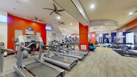 This single-floor fitness center is thoughtfully designed to accommodate a wide range of workout preferences and training styles. The space is visually energized by bold accent walls in vibrant red and deep blue, contrasting against clean white ceilings and neutral-toned carpet flooring throughout the main exercise areas.

The cardio zone is generously appointed with a row of modern treadmills and additional cardio machines, all reflected in large floor-to-ceiling mirrors that amplify the sense of space. Multiple ceiling fans provide consistent airflow across the workout floor, complemented by recessed lighting for a bright, welcoming atmosphere. A wall-mounted television keeps members entertained during cardio sessions.

The strength training section offers a comprehensive selection of cable machines, multi-station weight systems, and benches, providing options for both machine-based and free weight workouts. A dedicated functional training area features a polished hardwood floor, floor-to-ceiling mirrors, stability balls, a BOSU trainer, resistance bands, and a dumbbell rack — ideal for stretching, balance work, and group or personal training sessions.

A standout feature of this facility is the full-sized racquetball court, enclosed with white walls, regulation court markings on a classic hardwood floor, and a glass entry door that connects directly to the main gym floor.