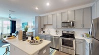 This single-floor residence offers a well-designed layout with modern finishes and a cohesive coastal-inspired color palette. The kitchen serves as a functional centerpiece, featuring gray shaker-style cabinetry extending to the ceiling, white subway tile backsplash, light quartz countertops, and a full suite of stainless steel appliances including a range, microwave, dishwasher, and refrigerator. A peninsula provides additional prep space and casual seating, while recessed lighting and wood-look plank flooring add to the polished aesthetic. The open layout connects the kitchen seamlessly to the living area, which benefits from natural light streaming through windows dressed with blinds and dark curtains. The bedroom is a standout space, anchored by a bold navy accent wall adorned with framed botanical artwork, a ceiling fan, and windows that invite natural light. The full bathroom features a tub-shower combination with striking navy and white striped curtains, a light-toned vanity with quartz countertops, and wood-look flooring. Throughout the residence, the consistent use of navy, white, and yellow accents creates a coordinated, inviting ambiance.