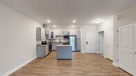 Located at 919 Woody Dr in Graham, NC, this single-floor unit presents a clean, contemporary design with thoughtful finishes throughout. The open-concept kitchen serves as the centerpiece of the living area, featuring gray shaker-style cabinetry, granite countertops, stainless steel appliances including a range with a stainless wall-mounted hood vent, built-in microwave, and refrigerator. A central kitchen island provides additional workspace and casual seating potential. Wood-look luxury vinyl plank flooring runs throughout the main living and kitchen areas, adding warmth and durability.

The bedrooms are carpeted for comfort and include ceiling fans with integrated lighting. Paired windows fitted with white blinds allow natural light to filter in while maintaining privacy. A spacious walk-in closet features wire shelving and hanging rods, offering generous storage capacity.

The bathrooms are well-appointed; one includes a walk-in glass-door shower, granite-topped vanity with gray cabinetry, and wood-look flooring, while another features a full tub-shower combination with tiled surround and built-in corner shelving. Additional storage is available via a dedicated pantry closet with wire shelving. The overall aesthetic is modern and move-in ready.