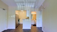 This single-floor residence offers a clean and functional layout designed to maximize comfort and usability. The open-concept kitchen serves as the heart of the space, outfitted with stainless steel appliances including a full-size refrigerator and range, crisp white cabinetry, and a peninsula breakfast bar that provides both additional prep space and a casual dining option. The kitchen's light color palette and efficient design create a welcoming culinary environment. Rich, dark hardwood-style flooring runs throughout, providing a striking contrast to the bright white walls and cabinetry and lending a warm, modern aesthetic to the interior. The layout flows naturally from the kitchen area toward the hallway, where a well-lit bathroom is conveniently accessible. The bathroom features white vanity cabinetry, a large mirror framed by a row of globe lights, and clean finishes throughout. A mirrored closet door along the hallway adds depth and reflects light, contributing to the overall sense of openness. The consistent use of white tones, modern fixtures, and cohesive flooring throughout the space creates a unified, contemporary atmosphere that is both practical and visually appealing.