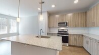 This well-appointed single-floor residence showcases a thoughtfully designed open-concept layout that seamlessly connects the kitchen and living areas. The kitchen serves as a central highlight, featuring a large island with a granite countertop, an undermount sink, and stylish pendant lighting overhead. Light wood-grain cabinetry lines the walls, complemented by stainless steel appliances including a gas range, over-the-range microwave, and refrigerator. A subway tile backsplash adds a clean, contemporary touch, while wood-look plank flooring extends throughout the main living areas, enhancing the sense of warmth and continuity.

The living space is open and airy, framed by neutral gray walls and recessed lighting, creating a versatile and inviting environment. The bedroom features plush wall-to-wall carpeting, blinds-equipped windows that allow ample natural light, and a ceiling fan for added comfort. The adjoining bathroom is finished with matching granite countertops, wood-tone cabinetry, an undermount sink, and a large framed mirror, maintaining a consistent design language throughout the home.

Additional practical features include a utility panel, ample storage including a walk-in closet area, and a front entry door with access to natural light. Overall, the space presents a clean, modern aesthetic with quality finishes throughout.