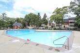 This single-story community clubhouse offers a variety of amenities designed for recreation and relaxation. At the heart of the outdoor area is a generously sized swimming pool with clear turquoise water, surrounded by a concrete deck, decorative fencing, and a small poolside utility structure. Mature trees frame the background, creating a pleasant, park-like atmosphere.

Inside, the clubhouse is organized into several distinct functional areas. A lounge and seating area features floor-to-ceiling windows that provide unobstructed views of the pool and landscaped grounds, filling the space with natural light. Comfortable chairs are arranged throughout, offering a welcoming spot to gather or unwind.

An adjacent game room accommodates a full-size ping pong table alongside additional seating, a wall-mounted television, and framed artwork. The room features vaulted ceilings and large windows, contributing to an open, airy feel.

A separate area serves as a fitness room, visible through large glass panels from the interior lounge. Exercise equipment is clearly visible within, providing a dedicated space for physical activity.

The clubhouse also includes a reception or entry area with warm wood accents, pendant lighting, and additional seating, rounding out this comprehensive community facility.
