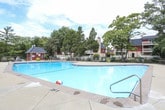 This single-story community clubhouse offers a variety of amenities designed for recreation and relaxation. At the heart of the outdoor area is a generously sized swimming pool with clear turquoise water, surrounded by a concrete deck, decorative fencing, and a small poolside utility structure. Mature trees frame the background, creating a pleasant, park-like atmosphere.

Inside, the clubhouse is organized into several distinct functional areas. A lounge and seating area features floor-to-ceiling windows that provide unobstructed views of the pool and landscaped grounds, filling the space with natural light. Comfortable chairs are arranged throughout, offering a welcoming spot to gather or unwind.

An adjacent game room accommodates a full-size ping pong table alongside additional seating, a wall-mounted television, and framed artwork. The room features vaulted ceilings and large windows, contributing to an open, airy feel.

A separate area serves as a fitness room, visible through large glass panels from the interior lounge. Exercise equipment is clearly visible within, providing a dedicated space for physical activity.

The clubhouse also includes a reception or entry area with warm wood accents, pendant lighting, and additional seating, rounding out this comprehensive community facility.