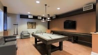 This single-floor entertainment space is thoughtfully designed for recreation and leisure. At the heart of the room sits a large, dark-stained pool table with a light felt surface, positioned beneath a pair of industrial-style pendant lights that provide focused illumination and add a modern aesthetic touch. Recessed ceiling lights complement the pendant fixtures, ensuring the space is well-lit throughout.

Adjacent to the pool table is a full-length shuffleboard table, offering additional gaming options. A wall-mounted flat-screen TV is prominently displayed on a warm-toned accent wall, creating a focal point for viewing and entertainment. Comfortable lounge chairs are arranged nearby, providing relaxed seating for spectators or those taking a break from the games.

The flooring throughout is a warm hardwood, contributing to the inviting and contemporary feel of the space. The walls feature a combination of neutral gray and earthy brown tones, giving the room a grounded, modern ambiance. Decorative artwork adds a personal touch to one of the walls, rounding out the interior design. HVAC vents are integrated into the ceiling and walls, ensuring climate comfort. Overall, this space is well-appointed and purpose-built for entertaining and social gatherings.